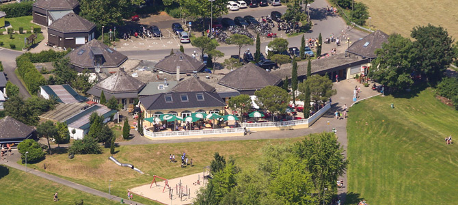 Luftaufnahme eines Parks mit einem Spielplatz, Sitzgelegenheiten im Freien mit Sonnenschirmen, umliegenden Gebäuden, Parkplatz und Grünflächen.