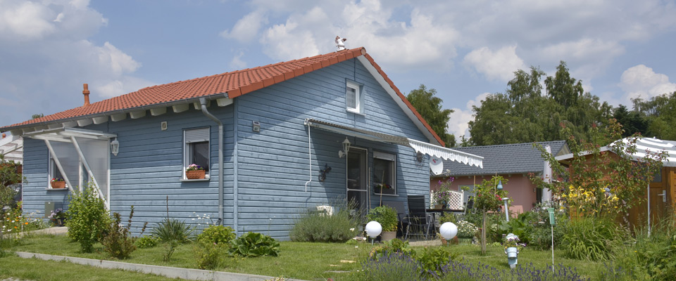 Ein kleines blaues Holzhaus mit einem roten Ziegeldach steht in einem Garten mit grünen Pflanzen und Blumen unter einem teilweise bewölkten Himmel.