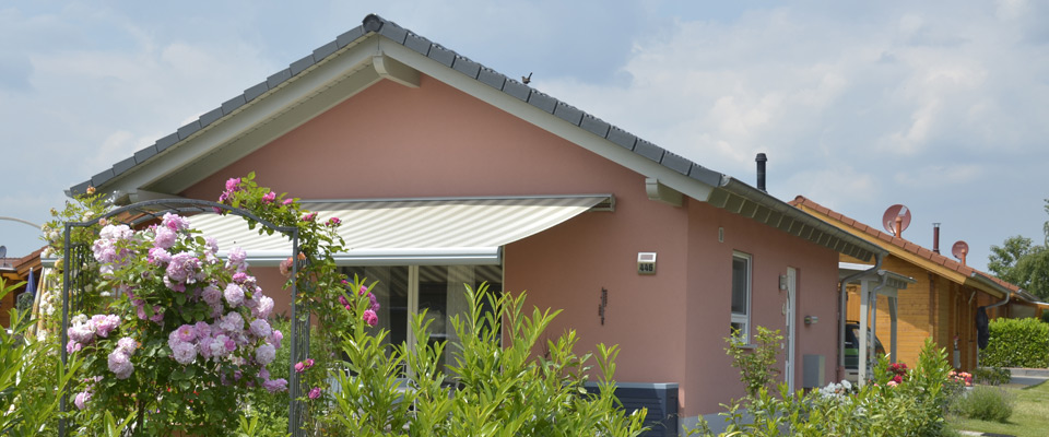 Einstöckiges Haus mit rosafarbenem Äußeren, weiß gestreifter, aufrollbarer Markise und Garten mit blühenden Blumen und Grünpflanzen, unter einem teilweise bewölkten Himmel.