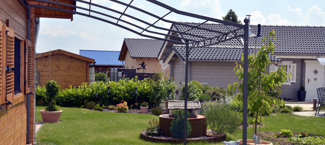 Ein Hinterhof mit einem grünen Rasen, Gartenbeeten, kleinen Bäumen, einem Holzschuppen und mehreren Häusern mit Ziegeldächern im Hintergrund.