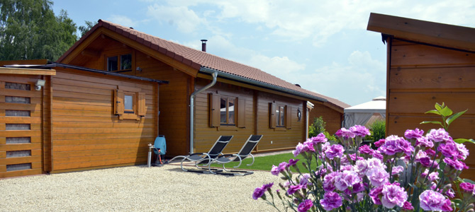 Ein Holzhaus mit rotem Dach steht neben einem Kiesweg und zwei Liegestühlen; im Vordergrund sind lila Blumen zu sehen.