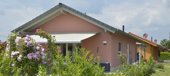 Einstöckiges rosafarbenes Haus mit gestreifter Markise, umgeben von grünen Sträuchern und blühenden rosafarbenen Blumen unter einem teilweise bewölkten Himmel.