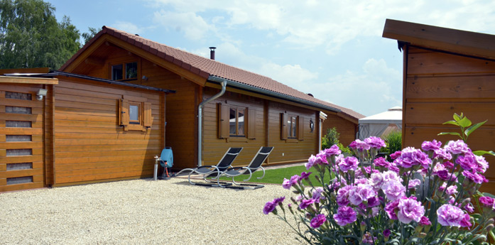 Ein Holzhaus mit zwei Liegestühlen auf einer Kiesfläche, umrahmt von Gras und blühenden rosa Blumen im Vordergrund.
