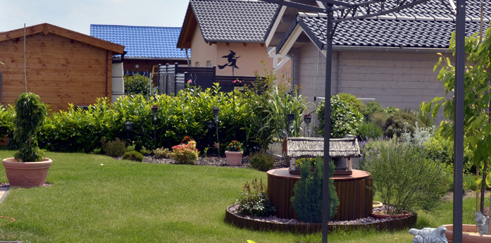 Ein gepflegter Garten im Hinterhof mit Topfpflanzen, einem kleinen Brunnen, gestutzten Sträuchern und benachbarten Häusern mit Ziegeldächern im Hintergrund.
