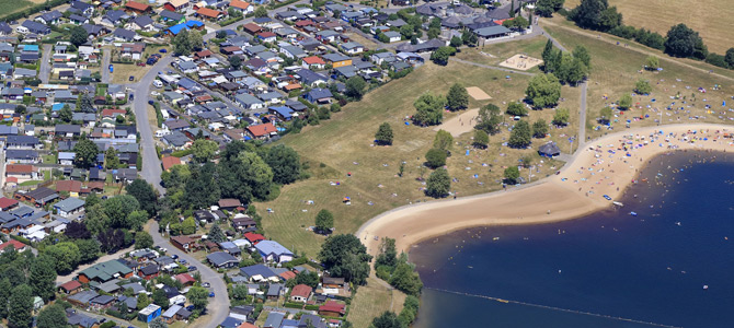 Luftaufnahme eines Wohnviertels neben einem Sandstrand mit verstreuten Menschen, grasbewachsenen Parkflächen und einem Gewässer.